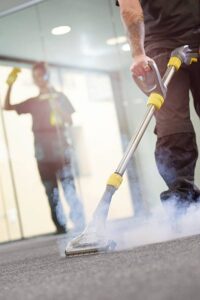 a male cleaning contractor steam cleans an office carpet in a empty office in between tenants. In the background a female colleague is polishing the glass partition offices .